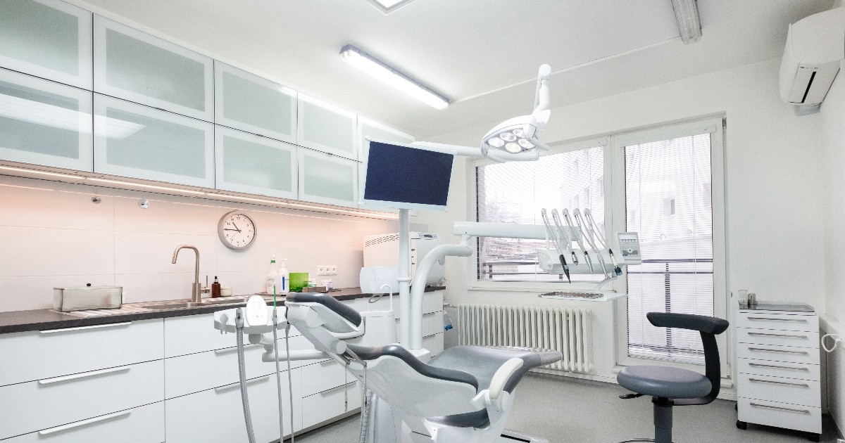 Imaginea prezintă interiorul unui cabinet stomatologic modern, dominat de culorile alb și gri, care include un scaun de tratament central, unitatea dentară aferentă și un ecran de monitorizare. Pe peretele din stânga se află un blat de lucru cu chiuvetă și dulapuri albe suspendate, iar lumina puternică, naturală și artificială, conferă spațiului o atmosferă de curățenie și sterilitate.