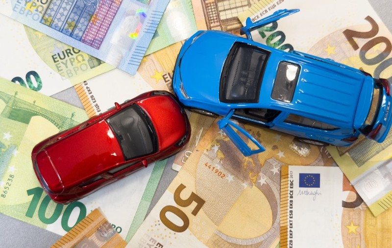 Două mașinuțe de jucărie - una roșie și una albastră - așezate deasupra unui strat de bancnote euro cu cupiuri de 50, 100 și 200 de euro.