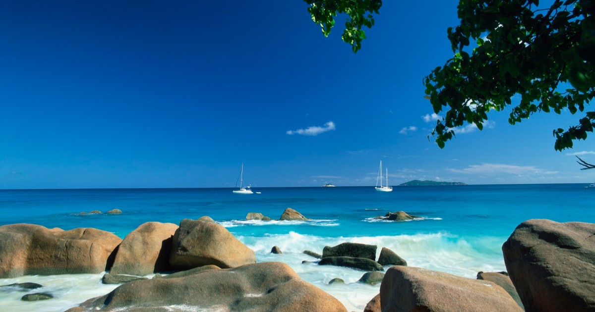 O imagine exotică de pe o plajă din insula Praslin, Seychelles. În fundalul fotografiei se observă iahturi ancorate sub un cer senin