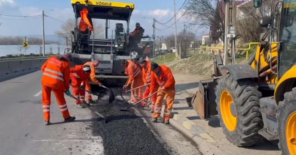 Mai mulți muncitori îmbrățai în costume portocalii lucrează la asfaltarea unei porțiuni de drum în Craiova, martie 2026
