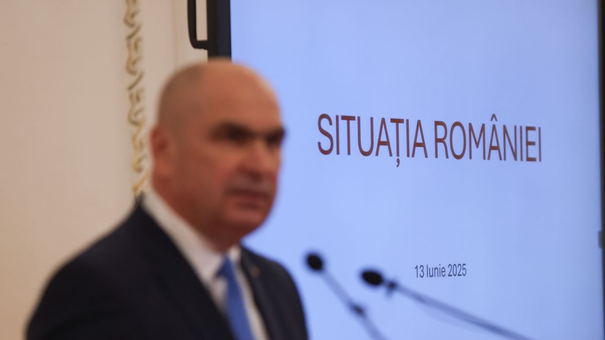 Imagine no sharp cu Ilie Bolojan in prim plan, in timpul unei conferinte, in fundal un panou pe care scrie Situatia României