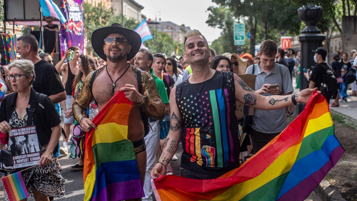 Imagine surprinsă în timpul Budapest Pride din iunie 2024. Doi bărbați se află în prim-plan, iar ambii țin în mâini câte un steag LGBTQ. Cel din stângă nu poartă tricou, are o pălărie și ochelari de soare. În spatele lor se află mai mulți oameni