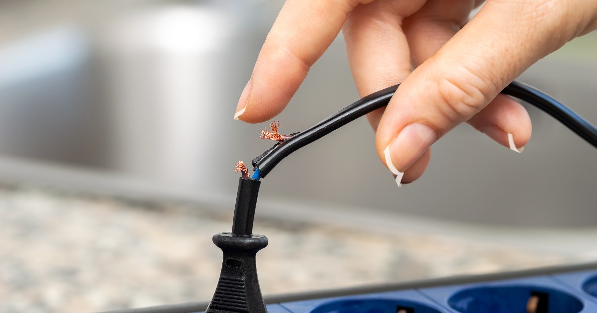 Imaginea surprinde o mână care atinge un cablu electric deteriorat, cu firele de cupru expuse, conectat la o prelungitoare. Fotografia evidențiază clar riscul de electrocutare cauzat de cablurile defecte și lipsa izolației corespunzătoare.