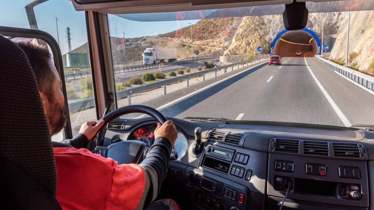Un șofer la volanul unui camion. Bărbatul poartă o bluză roșie și conduce pe o autostradă din Spania, drumul este liber