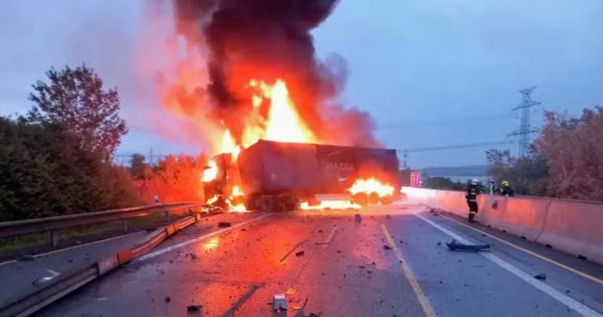Locul unui accident rutier pe o autostradă, unde un camion este complet cuprins de flăcări violente și fum negru, în timp ce pompierii intervin printre resturile împrăștiate pe carosabil.