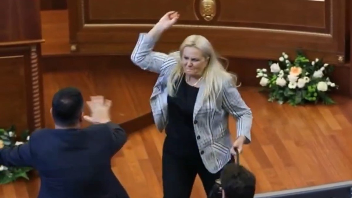 Imaginea prezintă o femeie blondă într-un sacou care ridică mâna pentru a arunca un obiect într-un bărbat dintr-o sală de parlament, în timp ce acesta încearcă să se apere.
