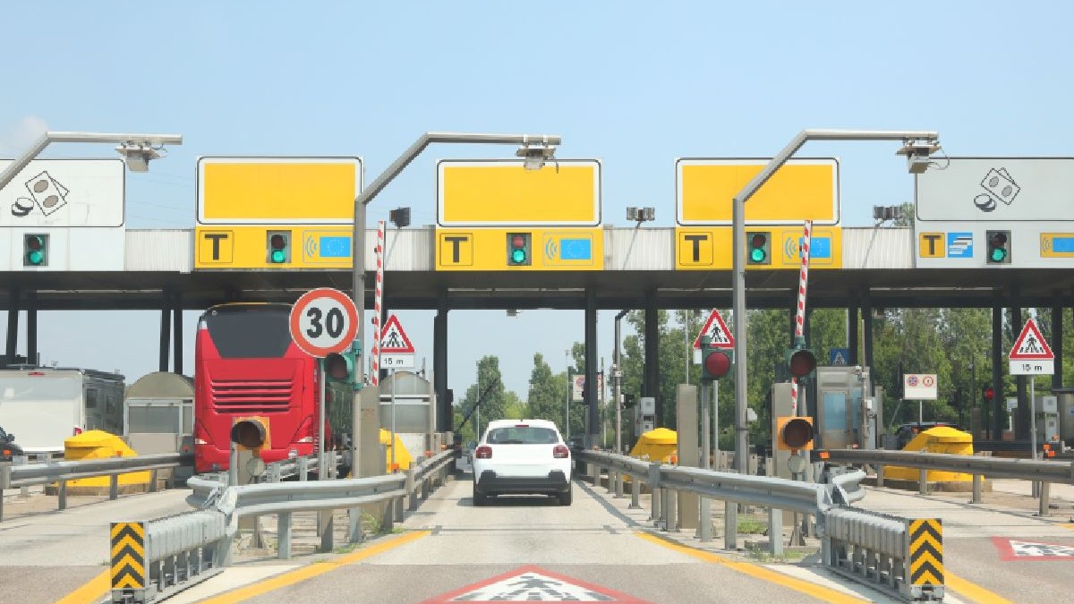 Fotografie cu mai multe cabine de taxare, indicatoare și semafoare, un autobuz roșu și o mașină albă oprite la bariere.