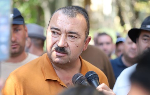 Un bărbat cu mustață vorbind în fața mai multor microfoane, înconjurat de oameni și jurnaliști