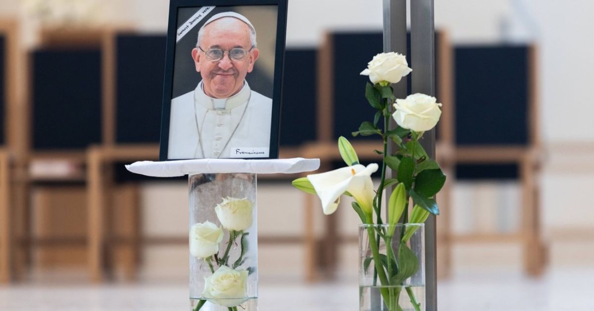 Papa Francisc a murit, imagine cu o fotografie cu Papa pe un pahar cu flori alve, alaturi un alt pagar cu trandafiri albi iar in spate niste scaune negre