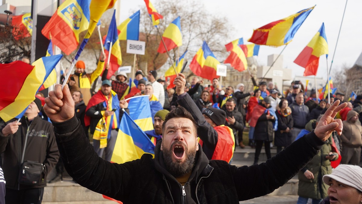Un bărbat susținător AUR țipă în timpul unui protest, ținând în mână steagul României. Alți oameni cu steaguri se văd în fundal