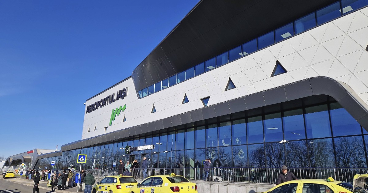 În imagine este surprins Aeroportul Internațional Iași, cu fațada modernă a terminalului și zona de sosiri, unde sunt parcate mai multe taxiuri. În fața clădirii se află pasageri și pietoni, sugerând un aeroport activ și un flux constant de călători.