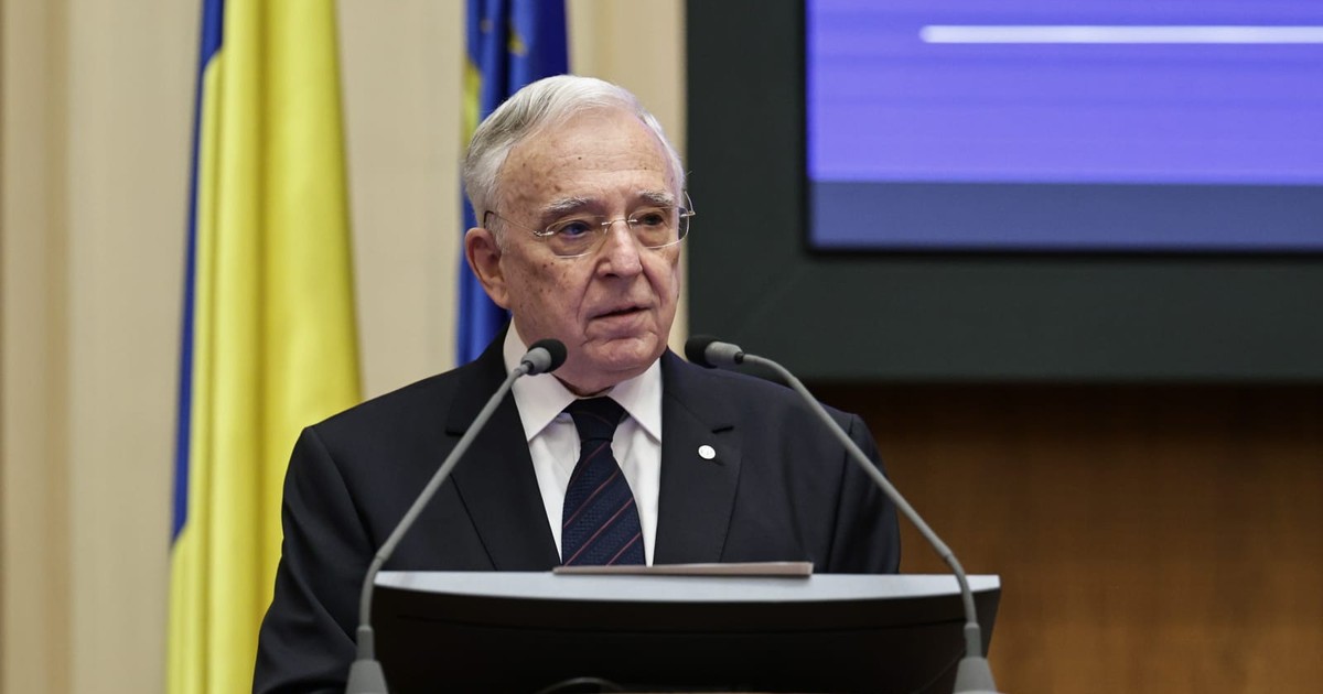 Mugur Isărescu, îmbrăcat într-un sacou negru, o cămașă albă și o cravată neagră, stând în spatele unui pupitru de lemn masiv prevăzut cu două microfoane, la o conferință de presă