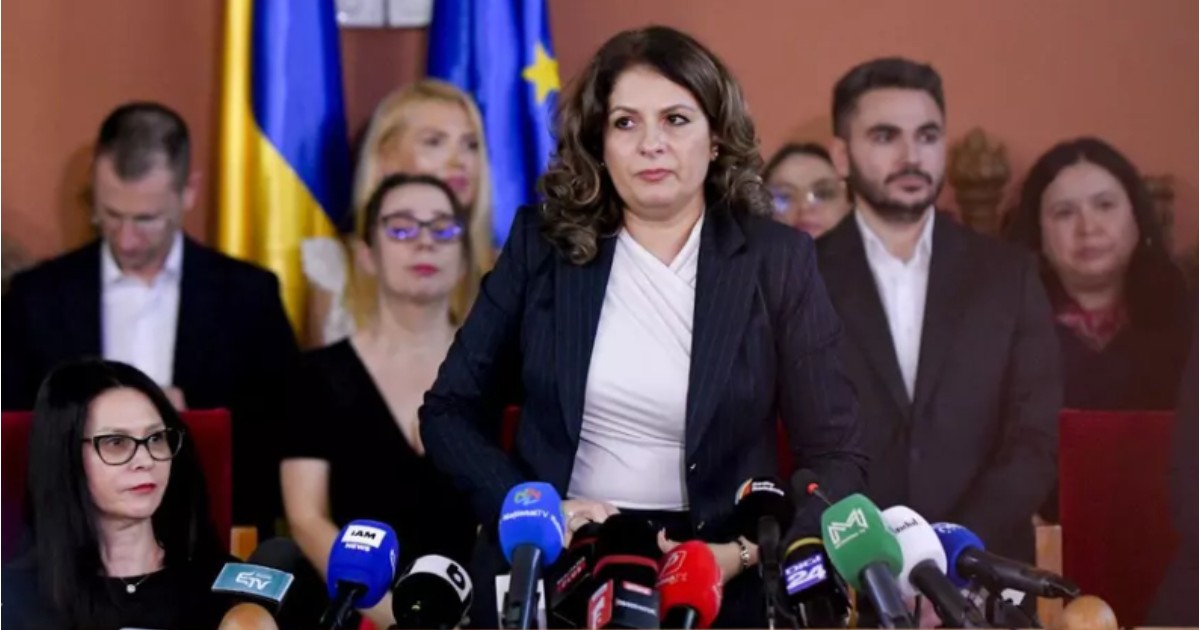 Liana Arsenie în timp ce stă în picioare în fața unui birou. În spatele său se află mai mulți oameni, în picioare.