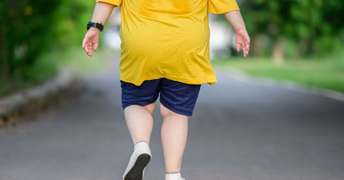Un copil gras fotografiat din spate, este îmbrăcat cu un tricou galben și blugi scurți, merge pe o stradă, dar nu i se vede capul