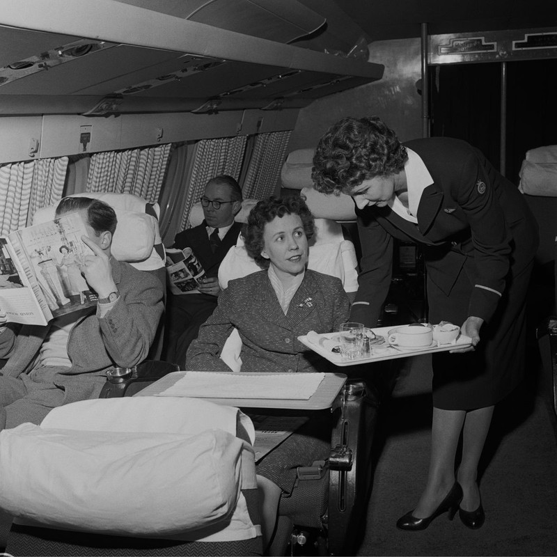 Însoțitoare de zbor pe avionul turbopropulsat Bristol Britannia, servește o tavă cu mâncare unui pasager în cabina avionului, 5 februarie 1957. Sursă foto: Getty Images