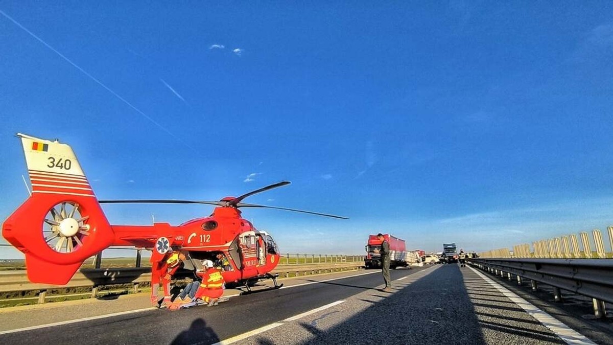 Un elicopter SMURD a aterizat pe autostradă, unde echipajele de intervenție acordă primul ajutor unei persoane rănite. În fundal se observă vehiculele implicate în accident și traficul oprit pe sensul de mers.