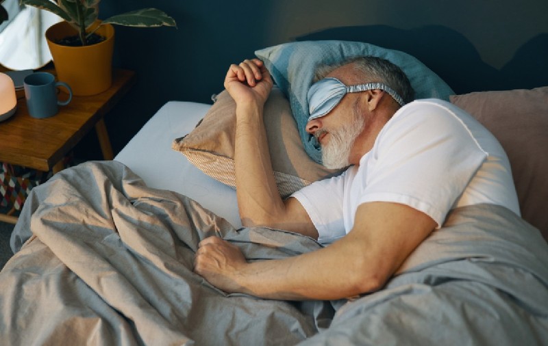 Om senior odihnindu-se în pat cu mască de somn, care apare relaxat și confortabil, înconjurat de perne și pături în dormitor confortabil