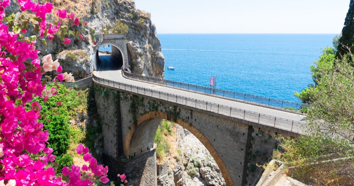 Podul de piatră de la Fiordo di Furore pe Coasta Amalfi, Italia, cu vedere spre marea albastră, flori roz în prim-plan și tunel în stâncă.