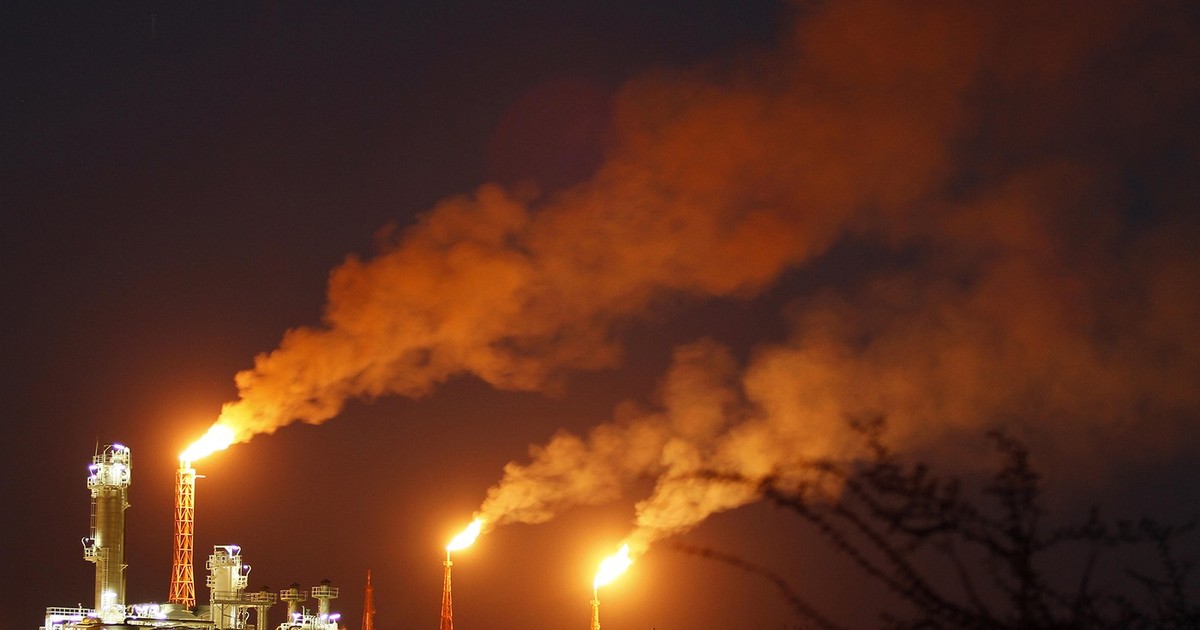 O fotografie de noapte care surprinde o rafinărie industrială în plină activitate, dominată de trei torțe uriașe din care izbucnesc flăcări puternice, luminând violent cerul întunecat și generând nori masivi de fum portocaliu, cu structurile metalice și conductele luminate artificial la bază, în timp ce în prim-plan se profilează conturul neclar al unor tufișuri.