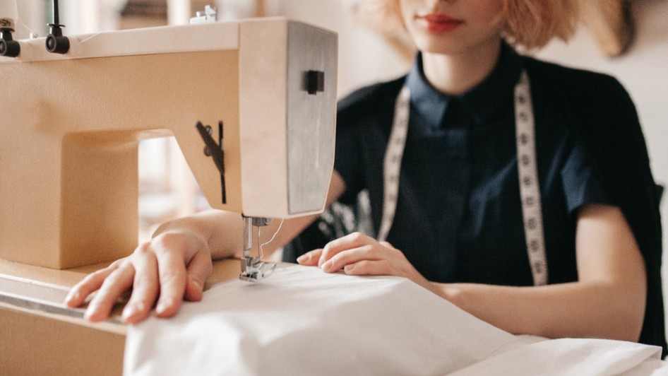 O croitoreasă cu un centimetru de croitorie în jurul gâtului, ghidând atent un material textil alb la o mașină de cusut electrică.