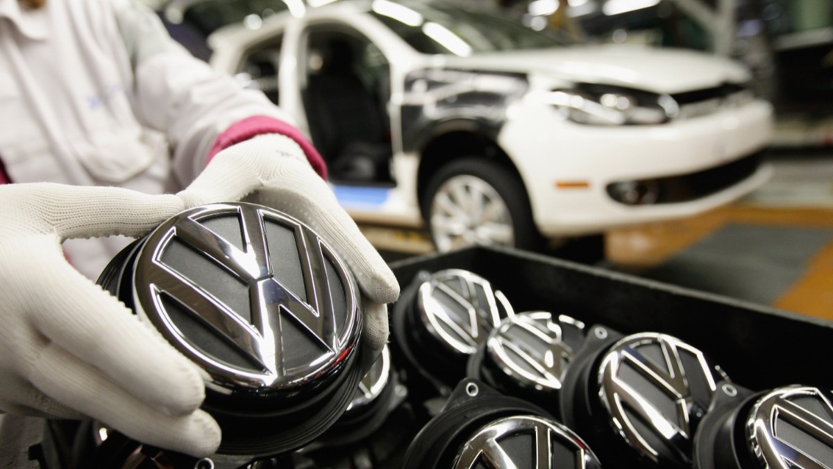 Imaginea prezintă un lucrător cu mănuși de protecție manevrând o emblemă cromatică Volkswagen pe linia de asamblare a unei fabrici auto.   