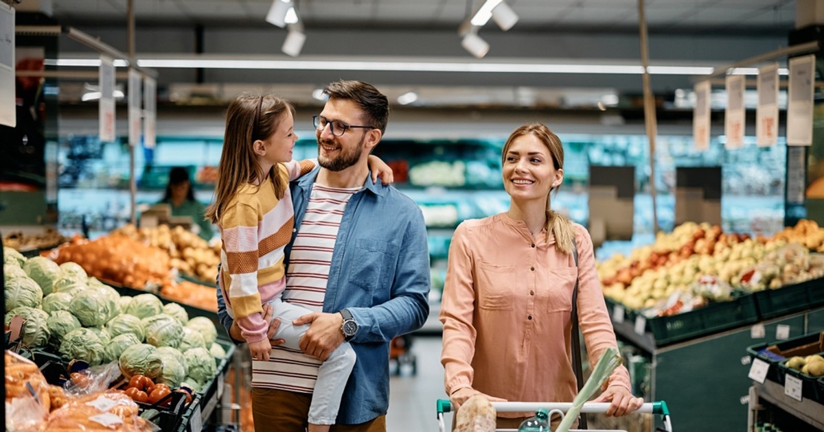 Program magazine de Crăciun 2023 - Imagine cu o familie, formată din părinţi tineri şi fiica lor, la cumpărături într-un supermarket.