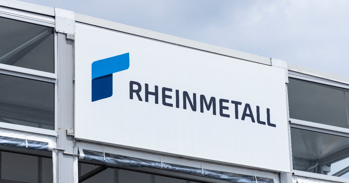 Imaginea prezintă o placă sau un panou publicitar montat pe structura exterioară a unei clădiri, afișând clar logo-ul și numele companiei "RHEINMETALL". Logo-ul este un simbol geometric, curbat, în nuanțe de albastru, poziționat în stânga numelui scris cu majuscule, sugerând sediul sau standul companiei la un eveniment.