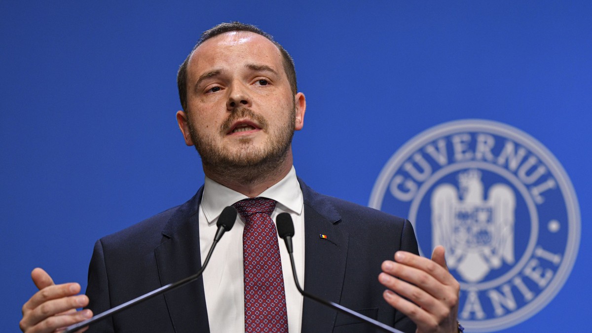 Alexandru Rogobete surprins în timpul unei conferințe de presă la Guvern vorbind și având mâinile ridicate. Poartă un sacou negru, o cămașă albă și o cravată grena