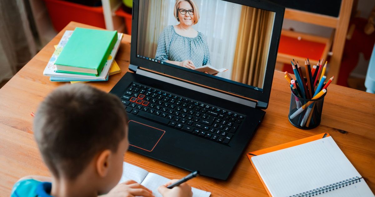 Un elev care stă cu spatele scrie pe caiet, iar în fața lui este un laptop deschis pe care comunică cu profesoara, care se vede pe ecran