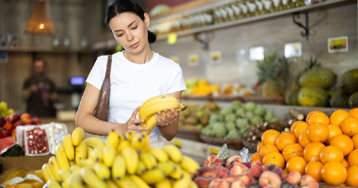 O femeie brunetă, îmbrăcată într-un tricou alb, cumpără banane proaspete într-un magazin, de la raionul de legume/fructe.