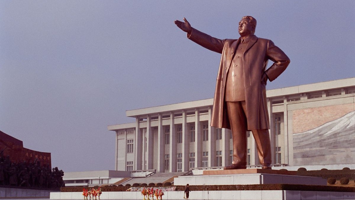 monument înalt, din material de culoarea bronzului, care îl înfățișează pe liderul nord-coreean Kim Jong-Un, cu o mână în șold și cealaltă mână întinsă în față, în fața unei clădiri albe cu coloane, Pyongyang, Coreea de Nord