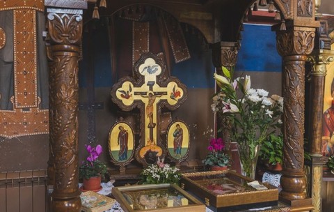 Cruce ortodoxă din lemn sculptat și pictat, așezată pe un altar decorat cu flori naturale într-o biserică.