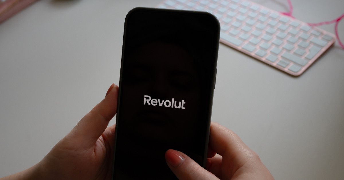 O persoană ține un smartphone negru în mâini, iar pe ecranul întunecat al acestuia se afișează logo-ul Revolut cu litere albe, semnalând deschiderea aplicației de banking. În fundalul neclar, se observă o tastatură de computer de culoare roz-pal, așezată pe o suprafață de birou deschisă la culoare.