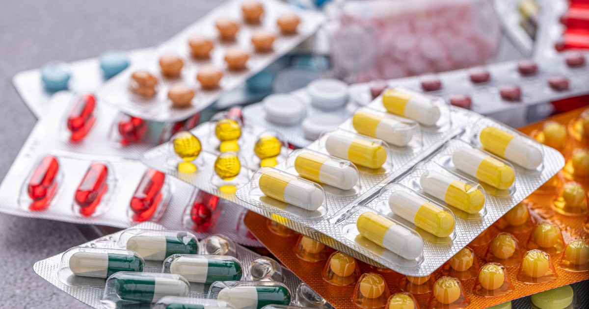 Imaginea prezintă un prim-plan al unei grămezi de diverse medicamente ambalate în blistere (folii) de culori diferite, incluzând pilule albe, capsule galben-albe și capsule verde-albe. Blisterele sunt așezate unele peste altele pe o suprafață gri, subliniind varietatea mare de pastile disponibile.