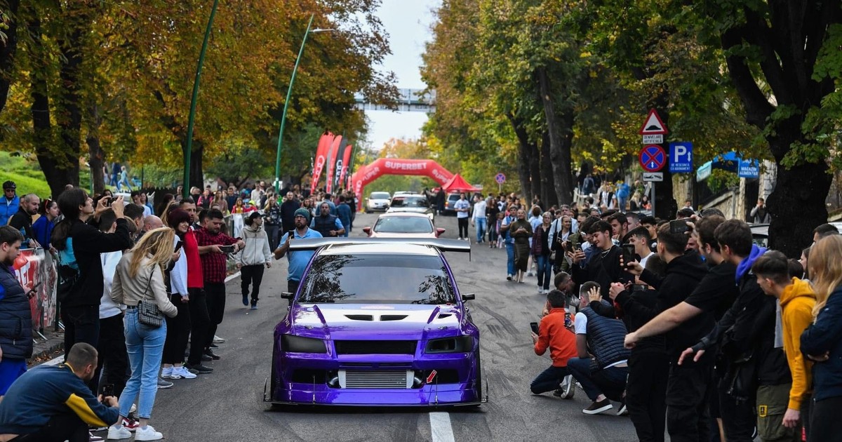 O mașină sport modificată, vopsită într-un violet intens, defilează pe o stradă plină de spectatori entuziasmați. Mulțimea o admiră și face poze