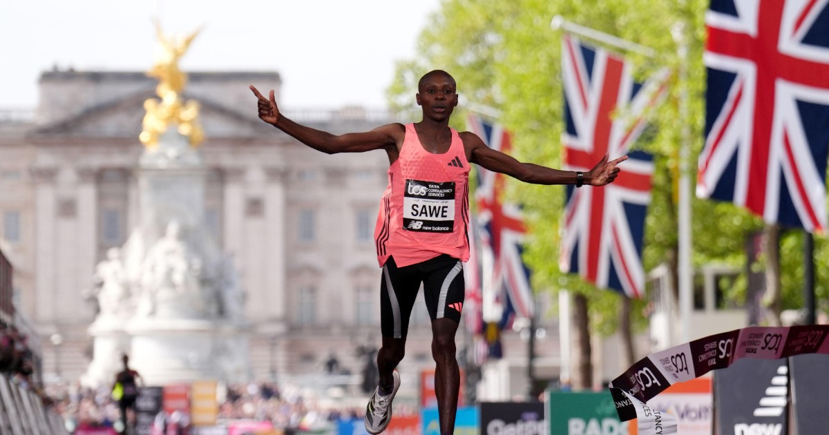 Un alergător kenyan trece linia de sosire cu brațele ridicate, celebrând victoria la maraton. În fundal se văd steaguri britanice și un reper iconic din Londra, sugerând atmosfera unei curse importante.