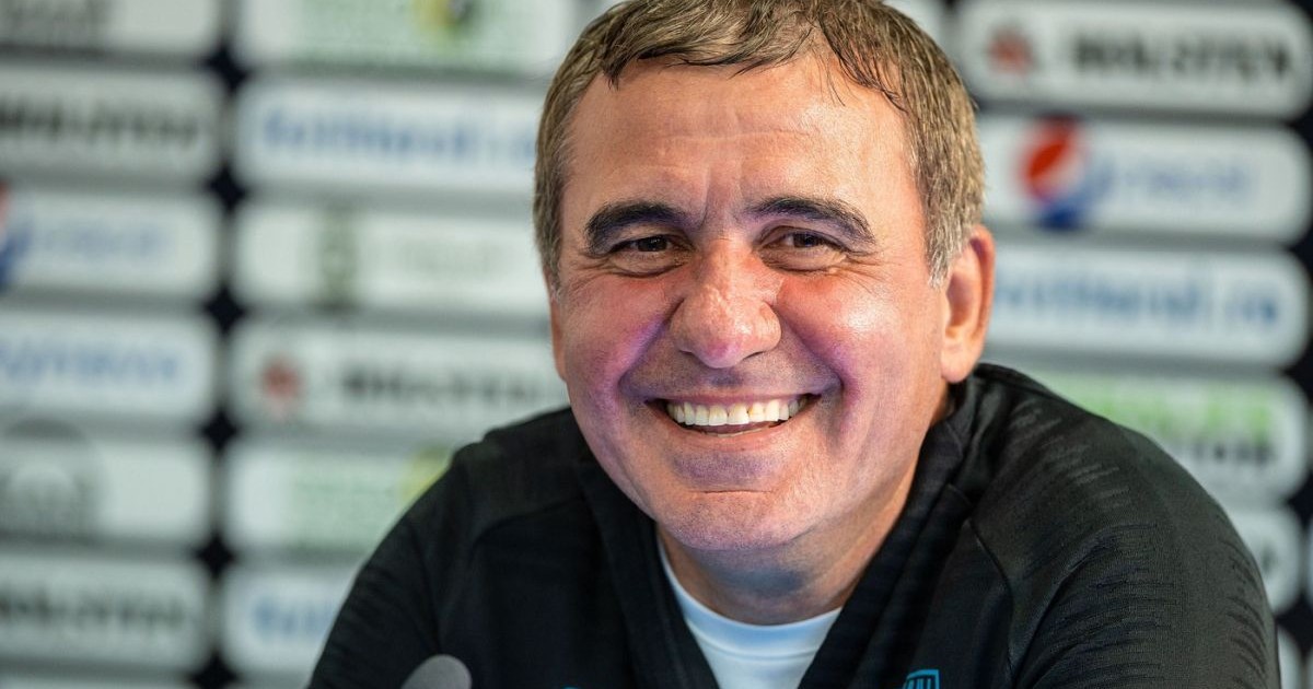 Gheorghe Hagi, îmbrăcat cu o bluză de trening neagră și tricou alb, râde, iar în fața lui este un microfon