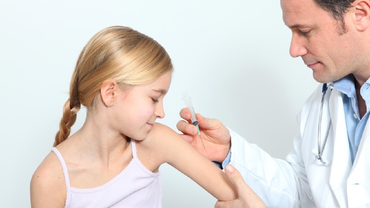 O fetiță blondă, cu părul prins în coadă, care primește o injecție în braț de la un medic îmbrăcat în halat alb, în timpul vaccinării.