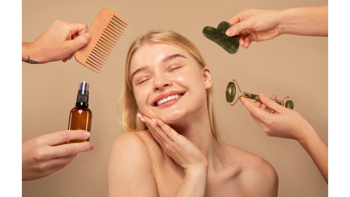Fata blonda care zambeste, In jurul ei sunt maini care intind un pieptan, un ser de fata si 2 accesorii pentru masaj facial