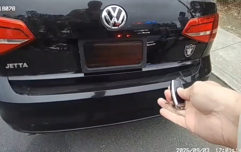 Partea din spate a unui autoturism Volkswagen Jetta negru, în timp ce o persoană acționează o telecomandă pentru a activa un dispozitiv ce acoperă numărul de înmatriculare.