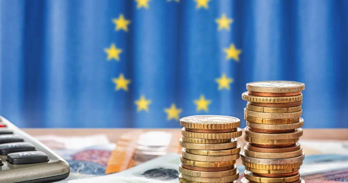 Monede așezate unele peste altele, bancnote, un calculator, iar în fundal este steagul Uniunii Europene
