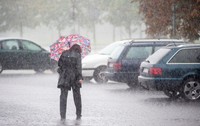 Imaginea prezintă o persoană îmbrăcată în haine închise la culoare care merge sub o umbrelă colorată în timpul unei ploi torențiale, într-o zonă de parcare cu autoturisme.   