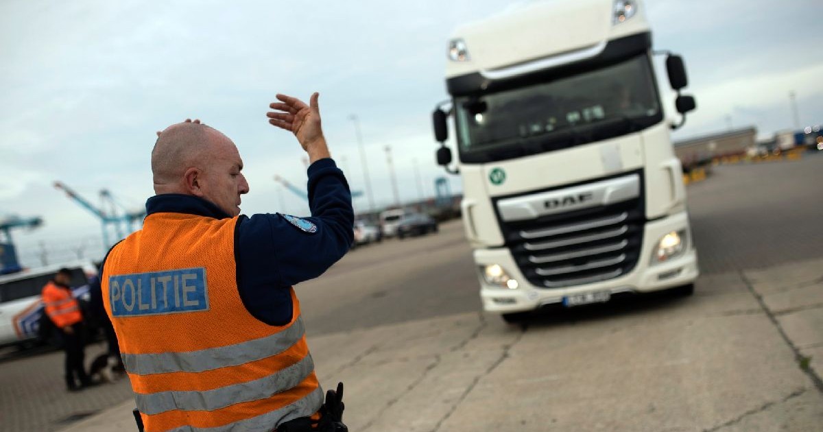 Un polițist îmbrăcat într-o vestă portocalie face semn cu mâna, dirijând un camion alb într-un port
