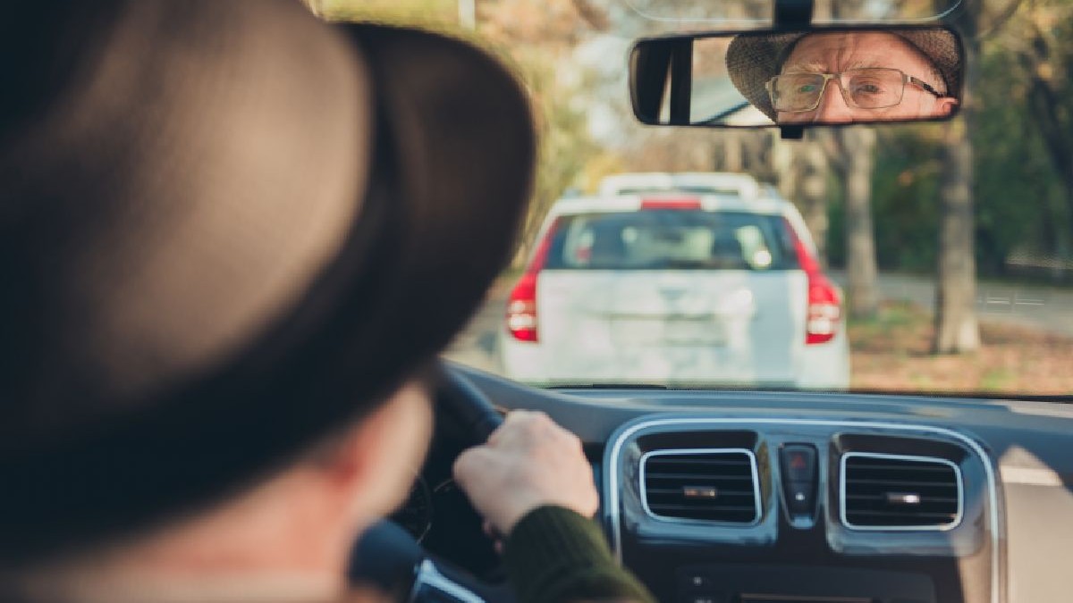 O fotografie arată o persoană cu pălărie maro și ochelari care se uită în oglinda retrovizoare, conducând o mașină cu un SUV alb în față.