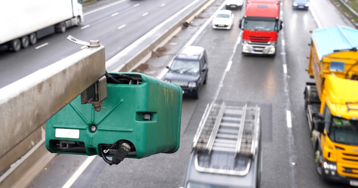 O cameră radar de culoare verde montată pe un suport metalic deasupra unei autostrăzi aglomerate, surprinzând în fundal mai multe camioane și autoturisme care circulă pe benzi.