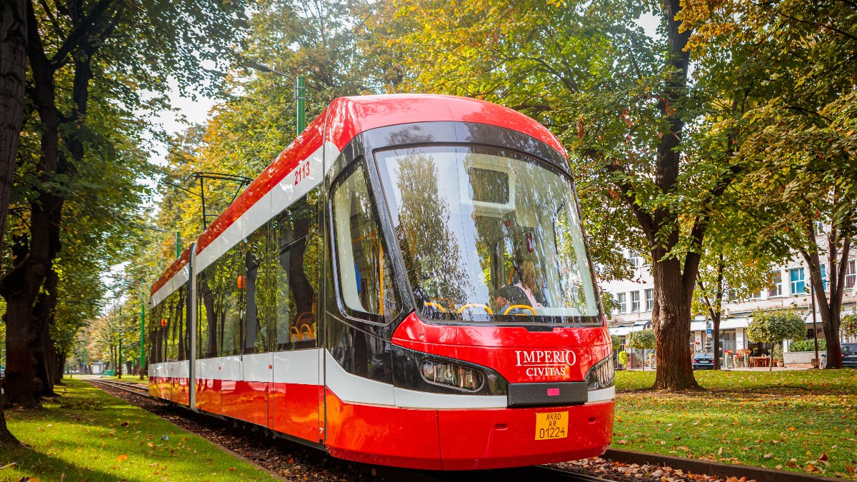 Imaginea prezintă un tramvai modern, de culoare roșie, care circulă pe o linie înconjurată de copaci și spațiu verde. Vehiculul este nou și bine întreținut, sugerând un sistem de transport public eficient și modern într-un oraș.