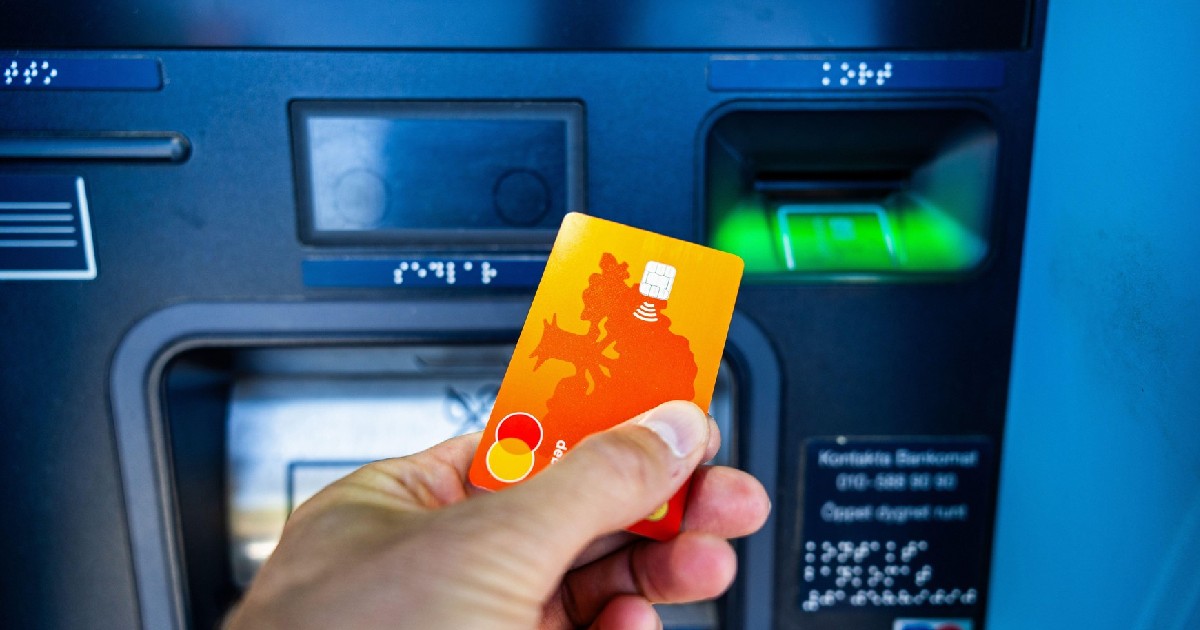 o mână ține un card portocaliu pe care urmează să-l introducă într-un bancomat