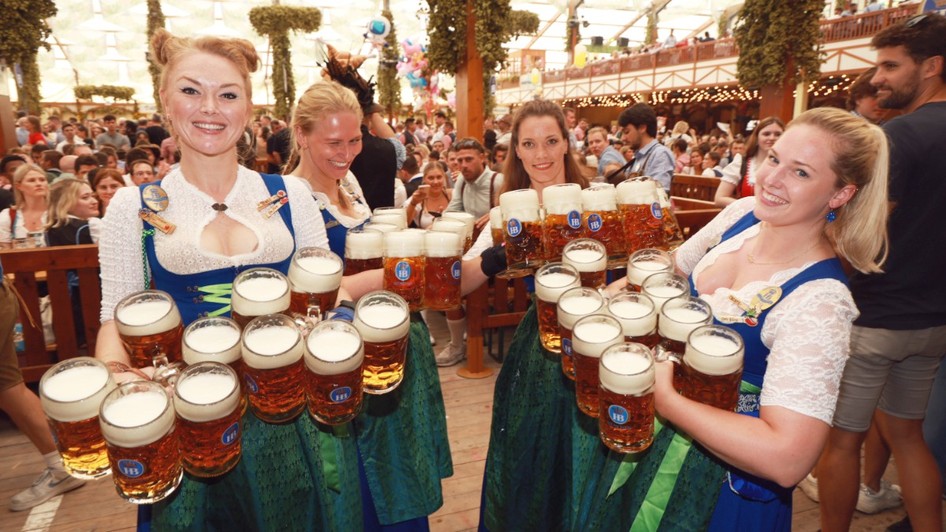 Mai multe chelnerițe îmbrăcate în costume tradiționale bavareze purtând numeroase halbe mari pline cu bere în interiorul unui cort aglomerat de la Oktoberfest.