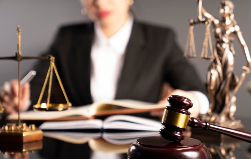 Imagine cu mai multe obiecte de pe biroul unui notar, o balanţă, o statuie a zeiţei Themis şi un ciocănel de justiţie, aflate pe un birou, în faţa unei femei
