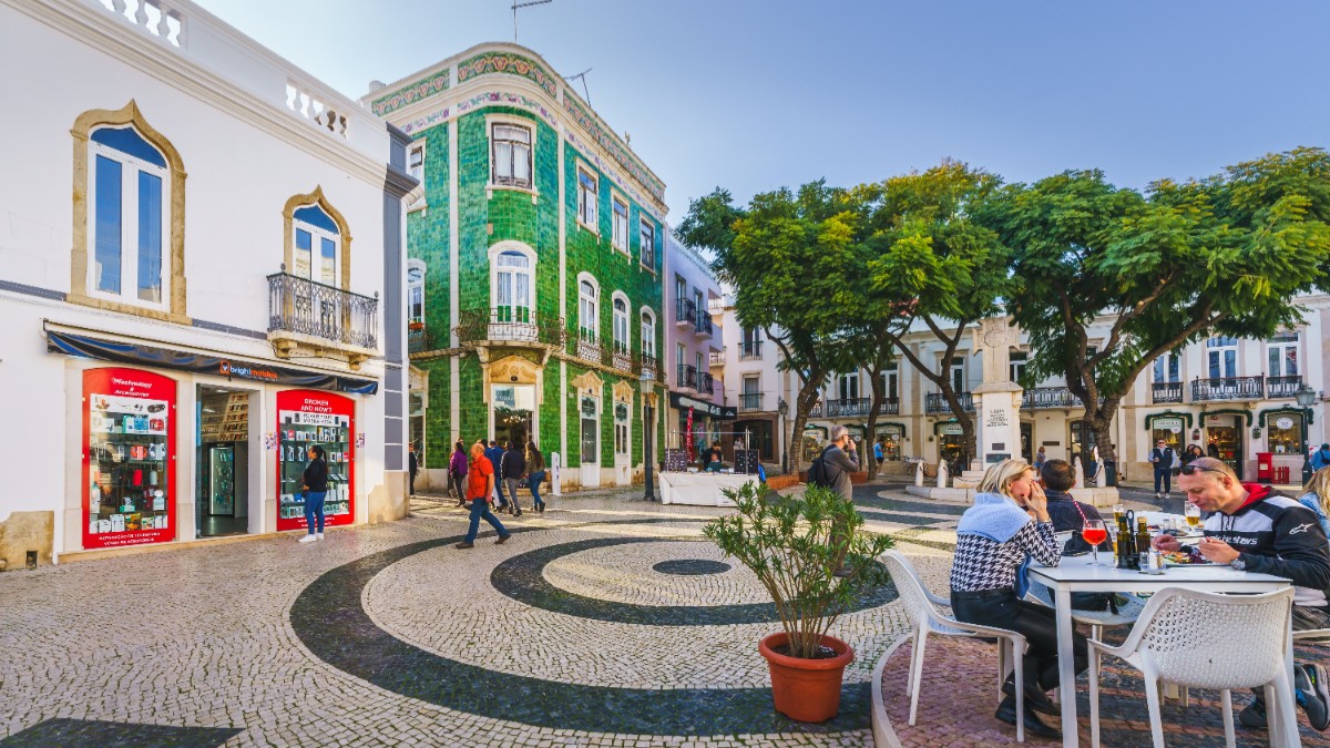 Un mic oraș din Portugalia cu o piațetă în centru, sunt case colorate în jur și mese albe cu scaune albe